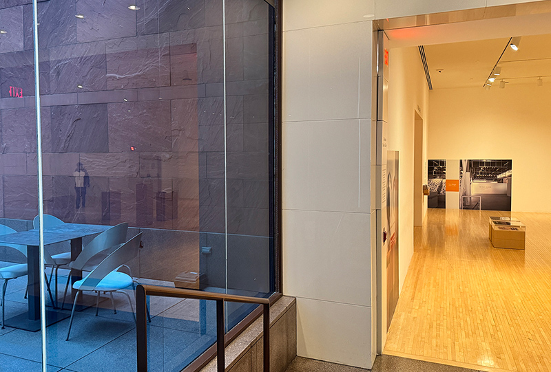 A view from the lobby of MOCA, the right half of the image looks through the doorway into the Michael Asher exibition, the left half of the image shows a table in the courtyard outside the museum, viewed through a glass window separating the courtyard from the museum lobby. Several pedestals and a museum guard are also visible, reflected in the window